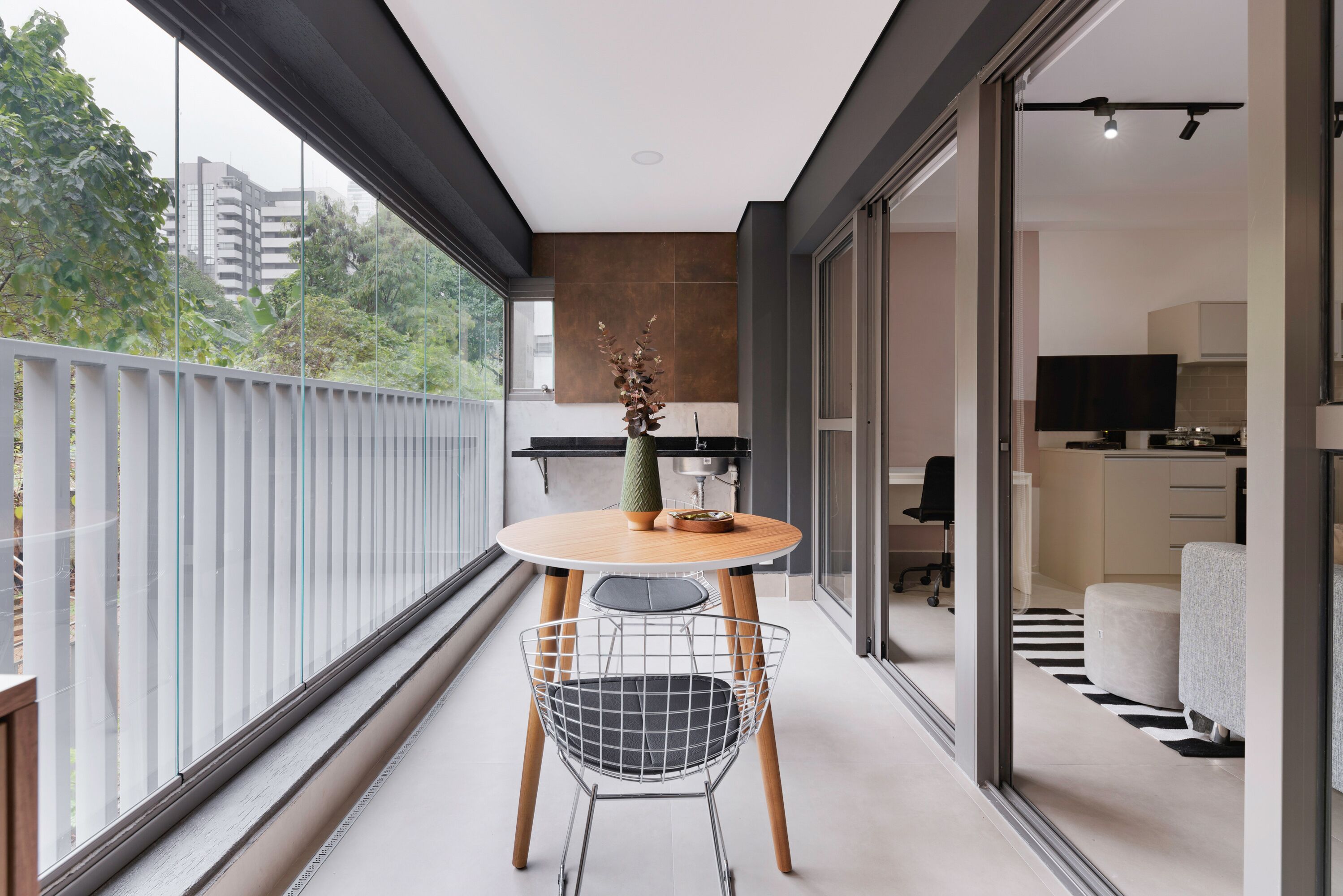 A modern balcony with a round table and wire chair, a view of a cityscape, and a glimpse of a kitchen area.