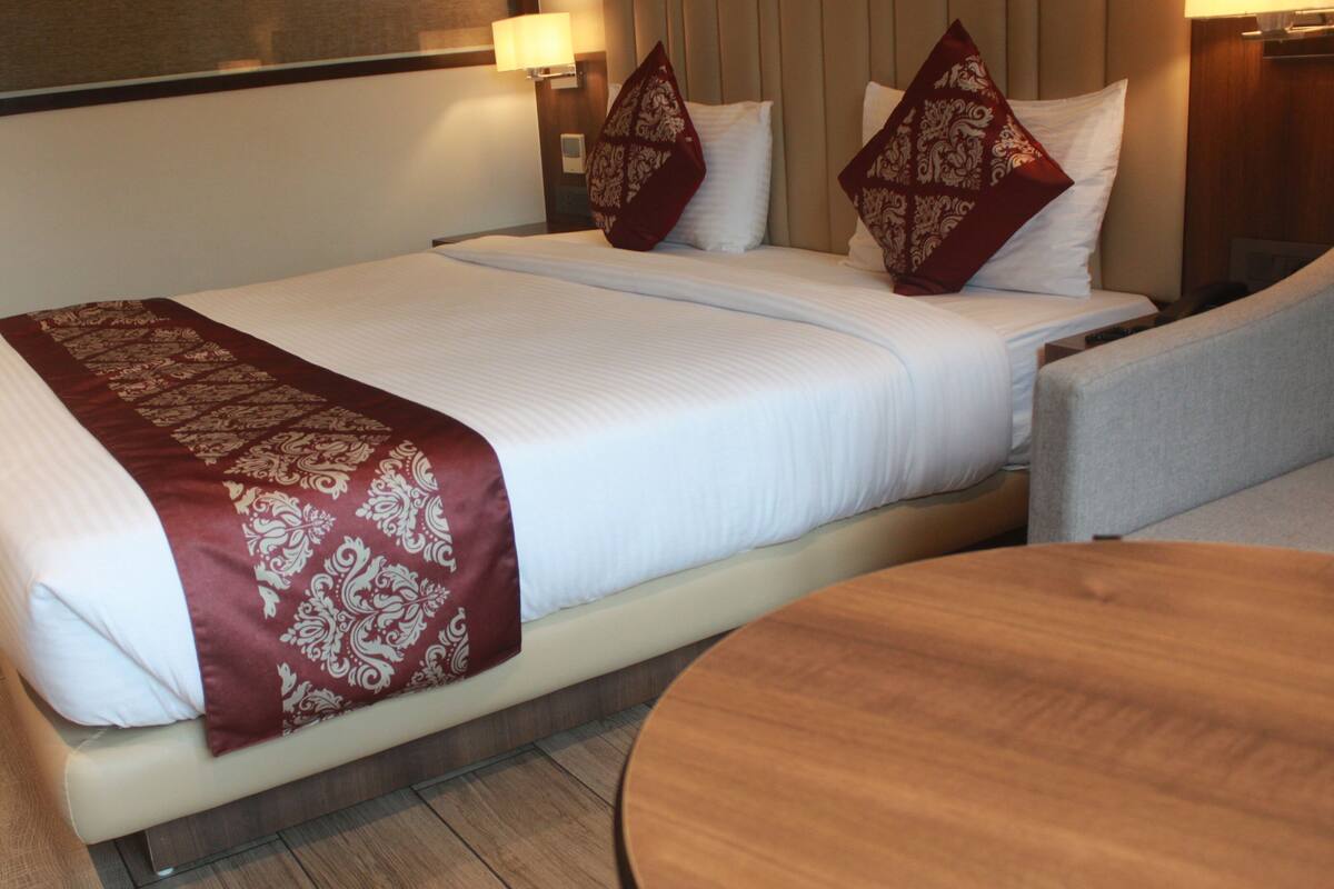 A hotel room with a bed, a wooden side table, and a wall-mounted lamp.