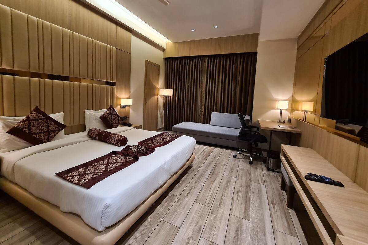 A hotel room with a large bed, a smaller sofa, a desk with a lamp, and a TV.