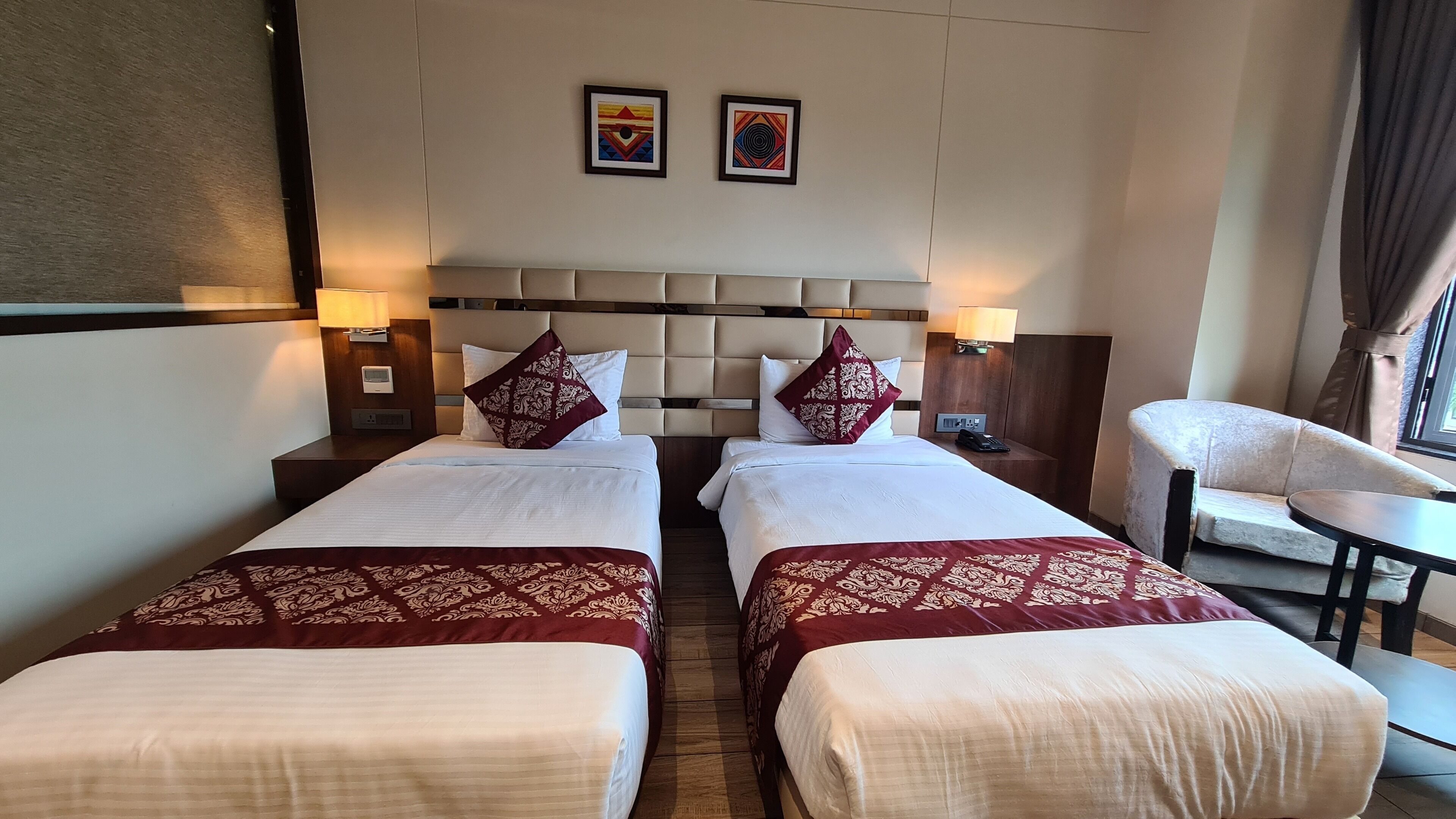 Habitación de hotel con dos camas, cada una con cabecera, mesita de noche con lámpara y una silla.