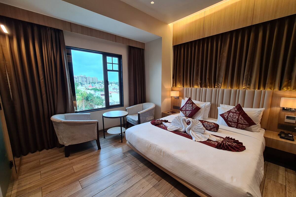 A hotel room with a large bed, a sitting area with a chair and table, a view of the city through a window, and a modern design with wooden paneling.