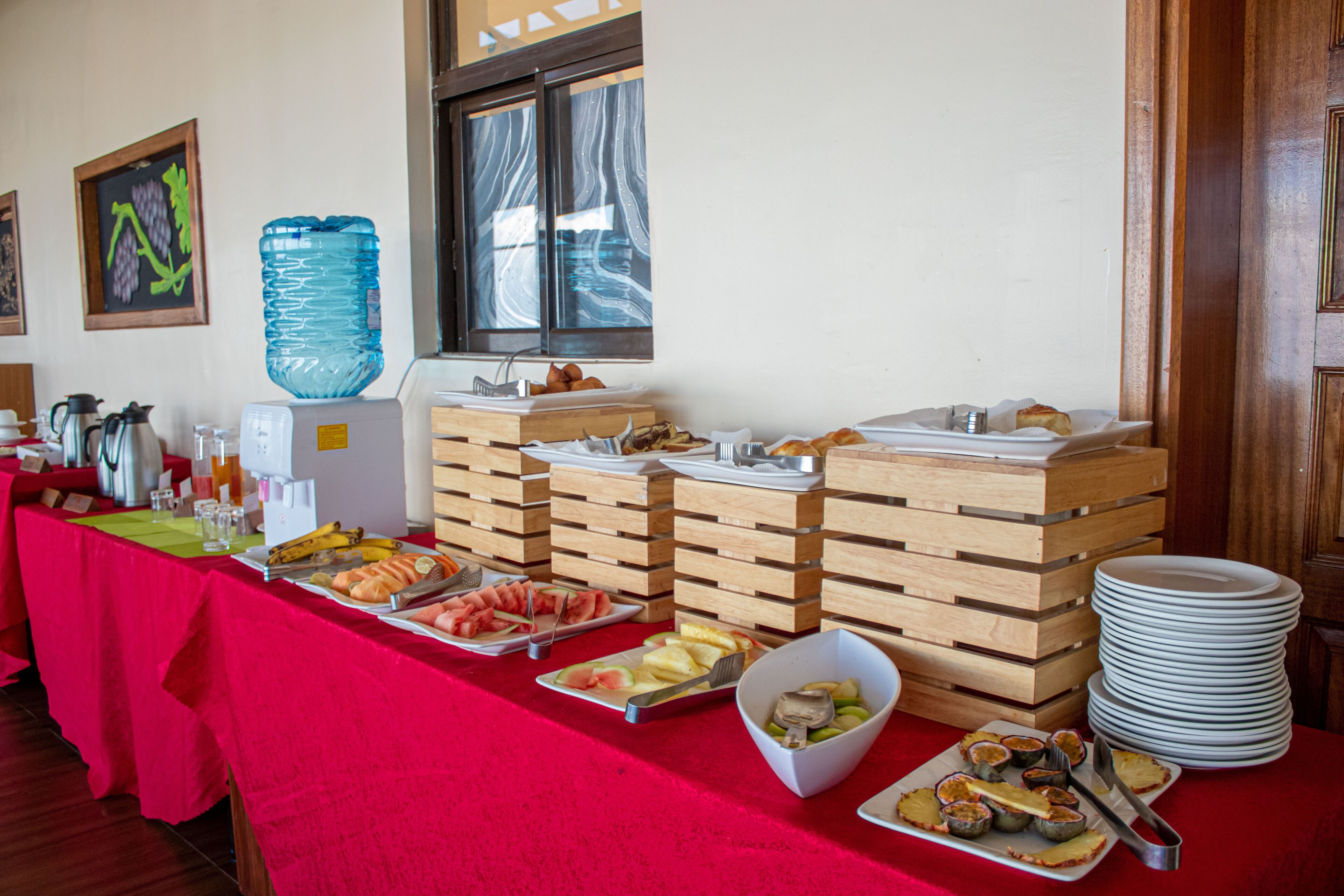 Montaje de buffet con diferentes alimentos, un dispensador de agua y platos apilados.