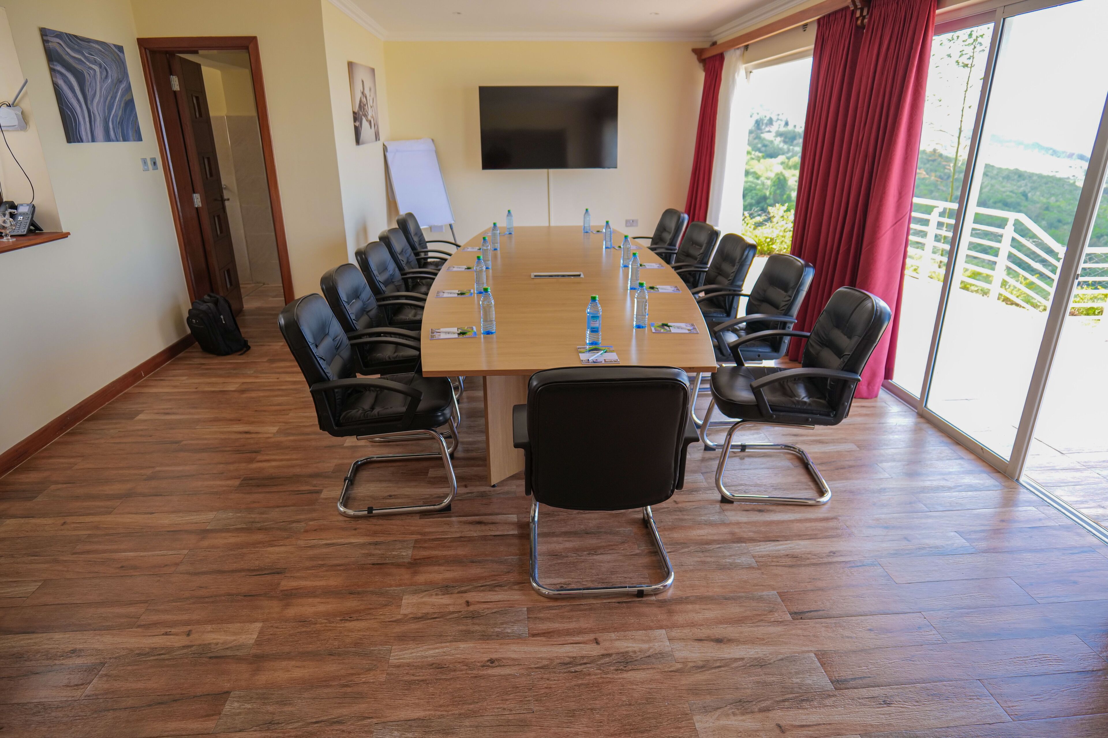 A conference room with a long table, black chairs, a TV on the wall, and a view of greenery outside.