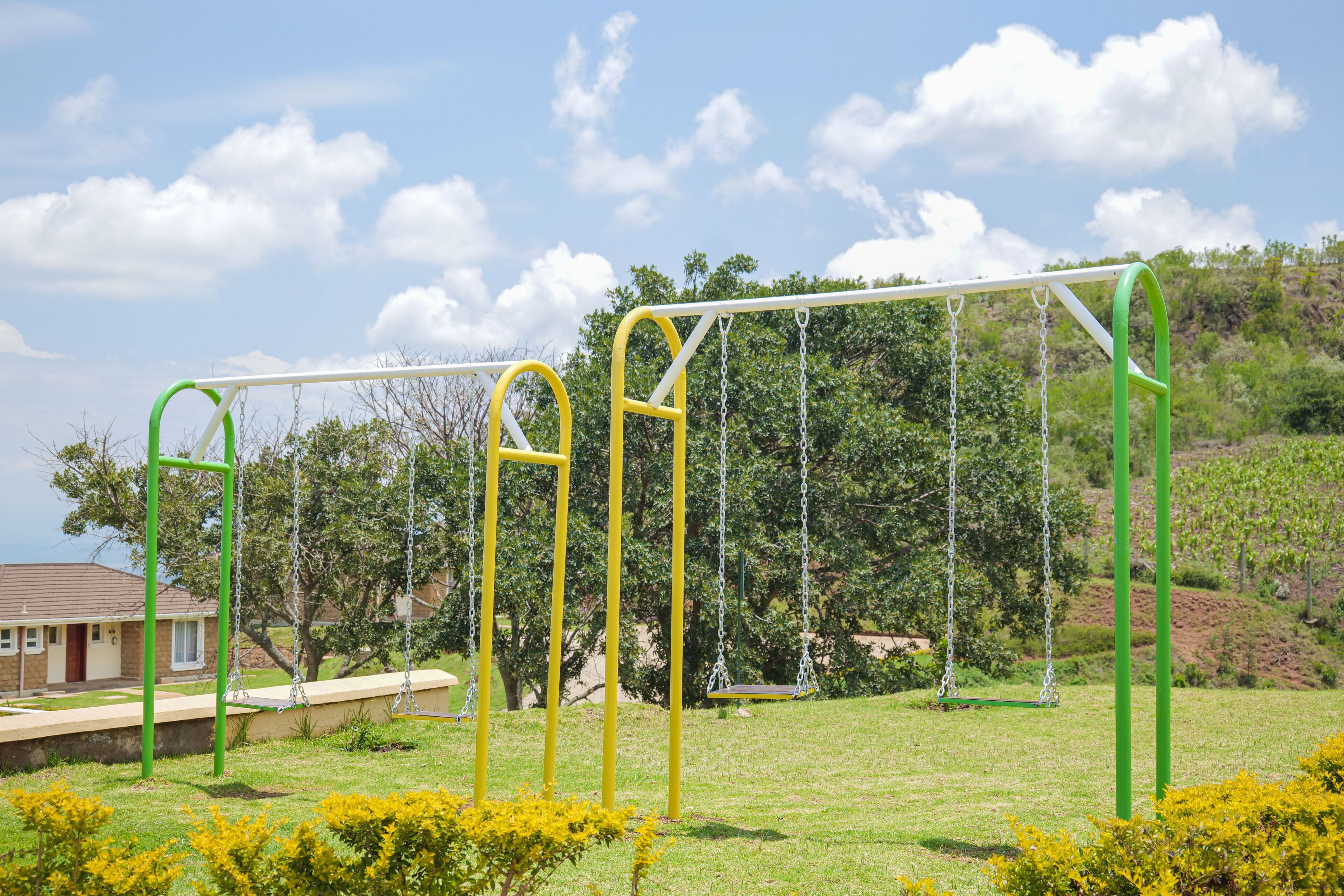 A playground with swings and a climbing frame in a grassy area with trees and a house in the background.
