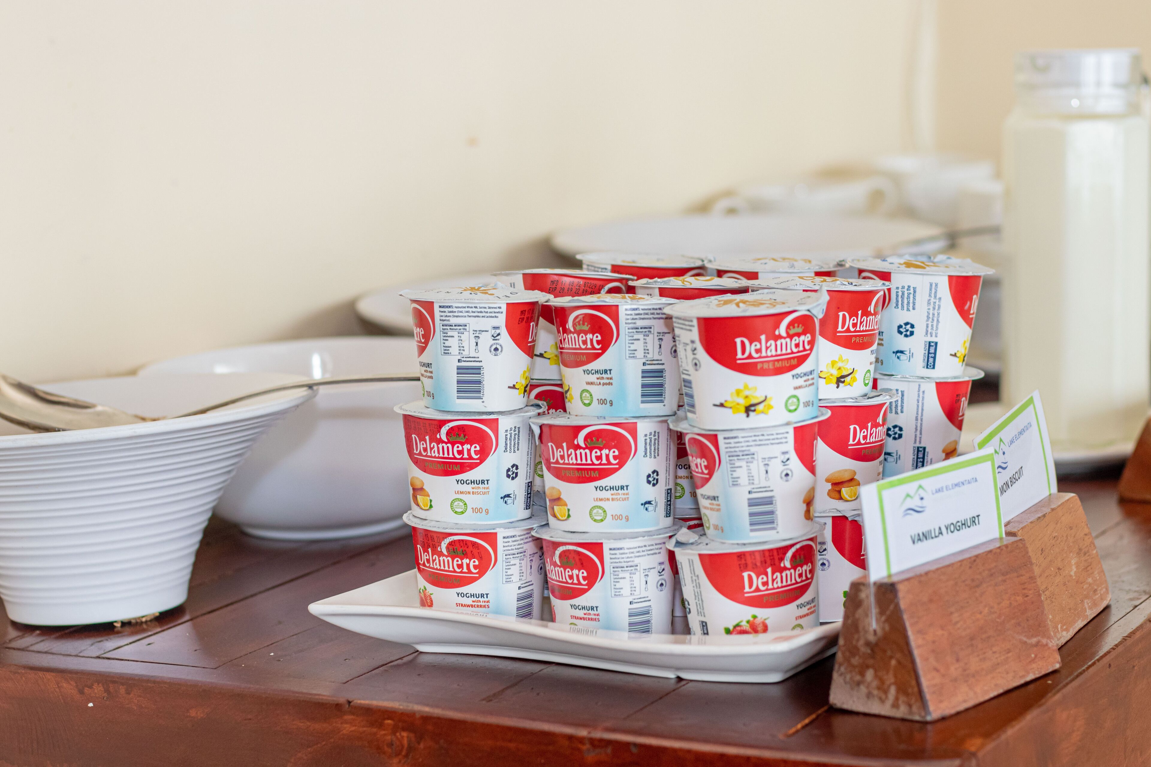 Exhibición de recipientes de yogurt Delamere apilados sobre una bandeja blanca, con etiquetas que señalan sabores como vainilla y fresa.