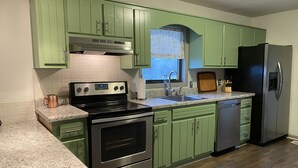 Una cocina con gabinetes verdes, electrodomésticos de acero inoxidable, encimera de granito y una ventana con cortina.