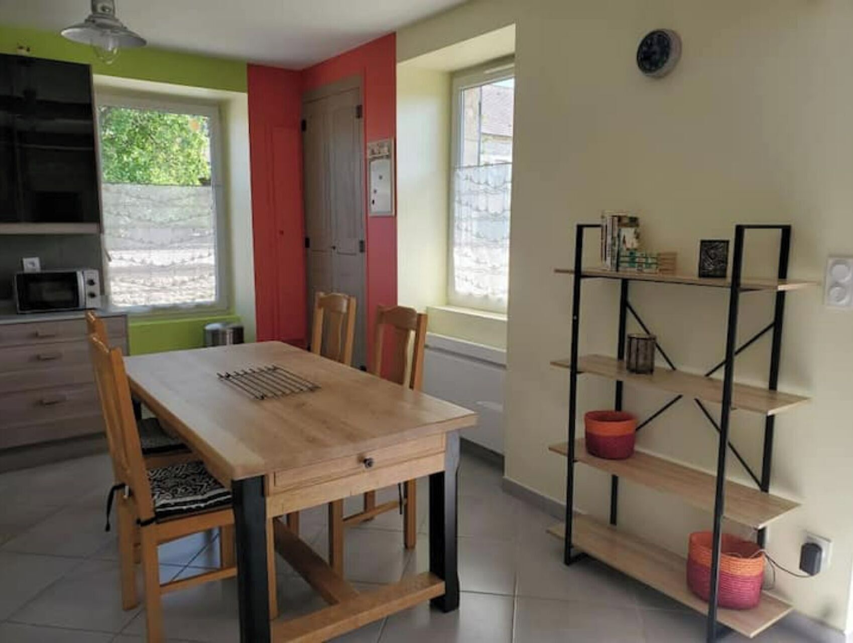 Un coin repas avec une table et des chaises en bois, une étagère en métal noir et une fenêtre donnant sur un paysage verdoyant.