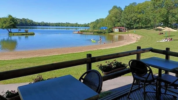 A lakeside view from a deck with tables and chairs, a grassy area with a small building, and a clear blue sky. - The fountains (Laives)