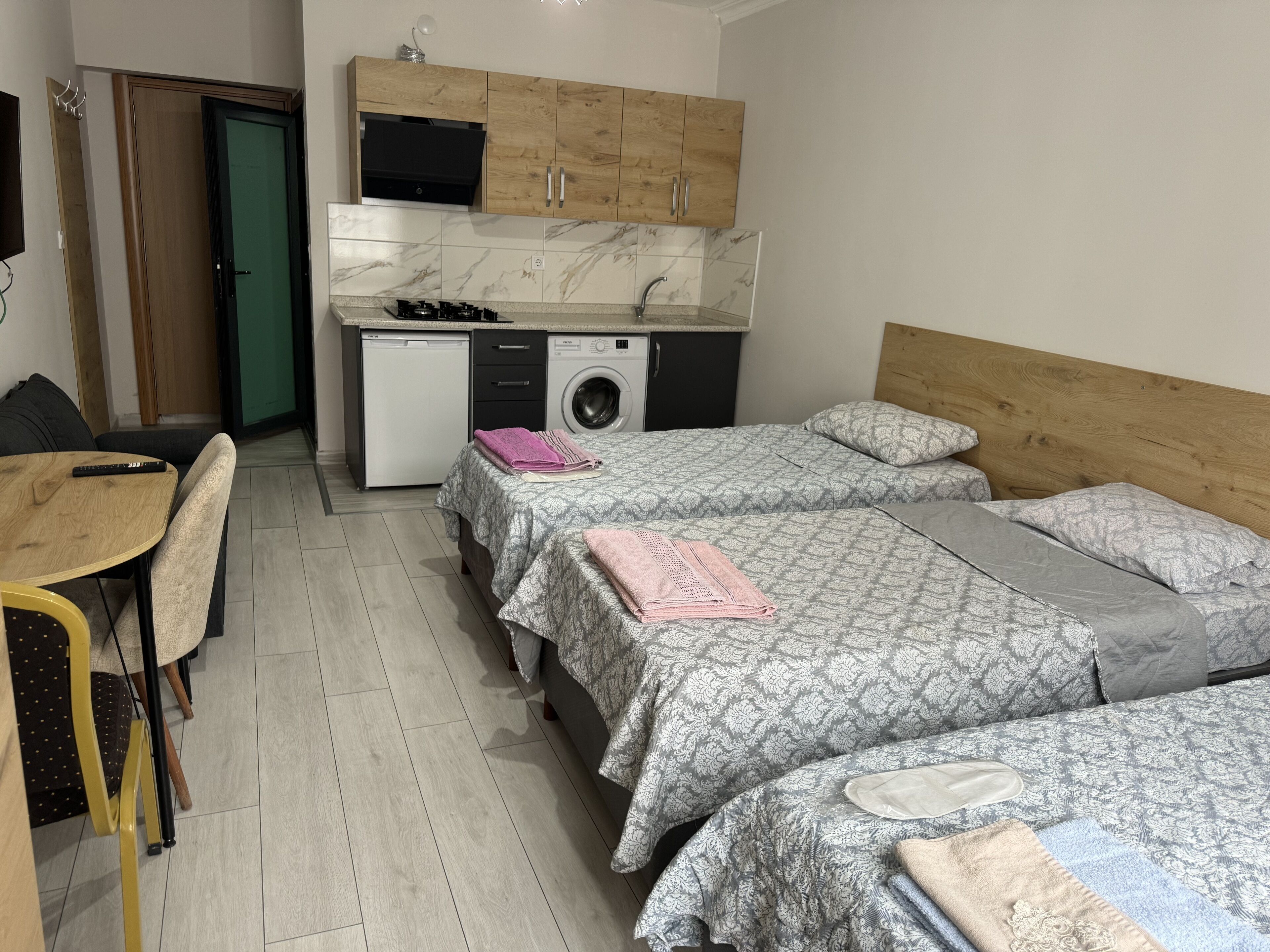 A compact bedroom with two beds, a small kitchenette, a washing machine, and a dining area with a table and chairs.