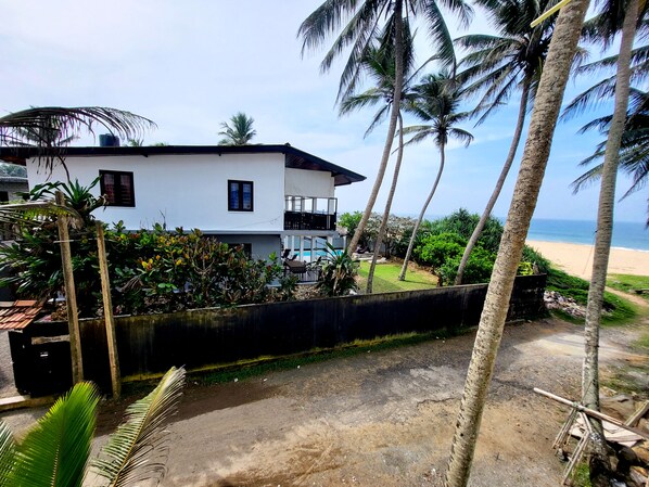 A white two-story house with a balcony, surrounded by palm trees and a view of the beach.