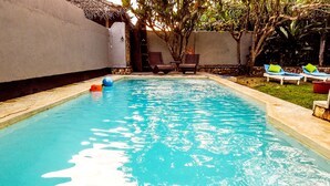 Une piscine rectangulaire à l’eau d’un bleu limpide, entourée d’un petit muret en pierre et d’une zone herbeuse agrémentée de chaises longues et d’une structure à toit de chaume.
