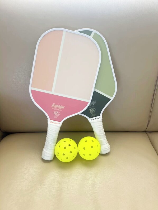 Two pickleball paddles with bright yellow balls on a beige leather surface.