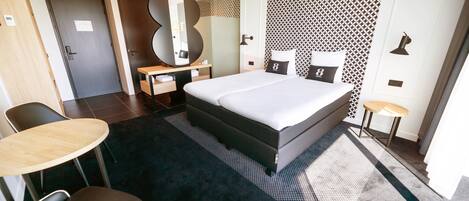 A modern hotel room with a large bed, a desk with a chair, a round table, and a mirror.