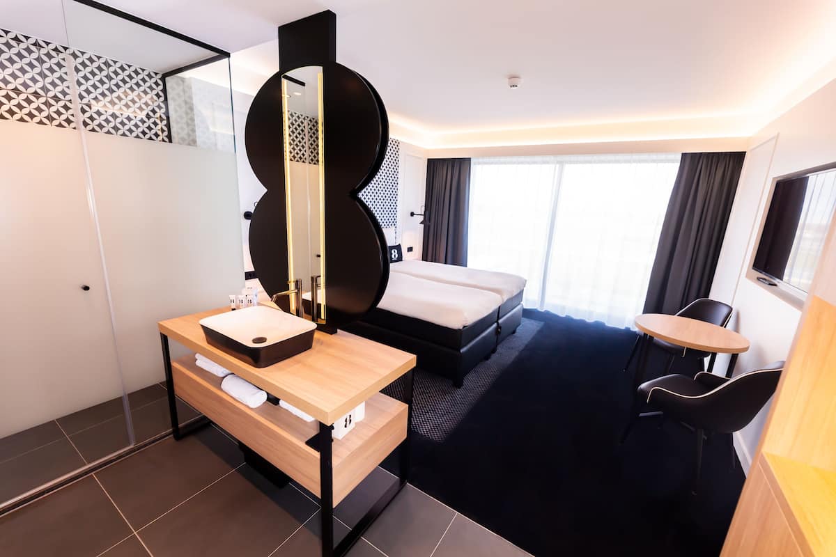 A modern hotel room with a bed, a bedside table with a mirror, a small desk, a chair, and a TV.