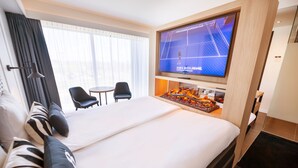 Habitación de hotel con una cama grande, un televisor de pantalla plana montado encima, una mesita pequeña, dos sillas y vistas a la ciudad por la ventana.