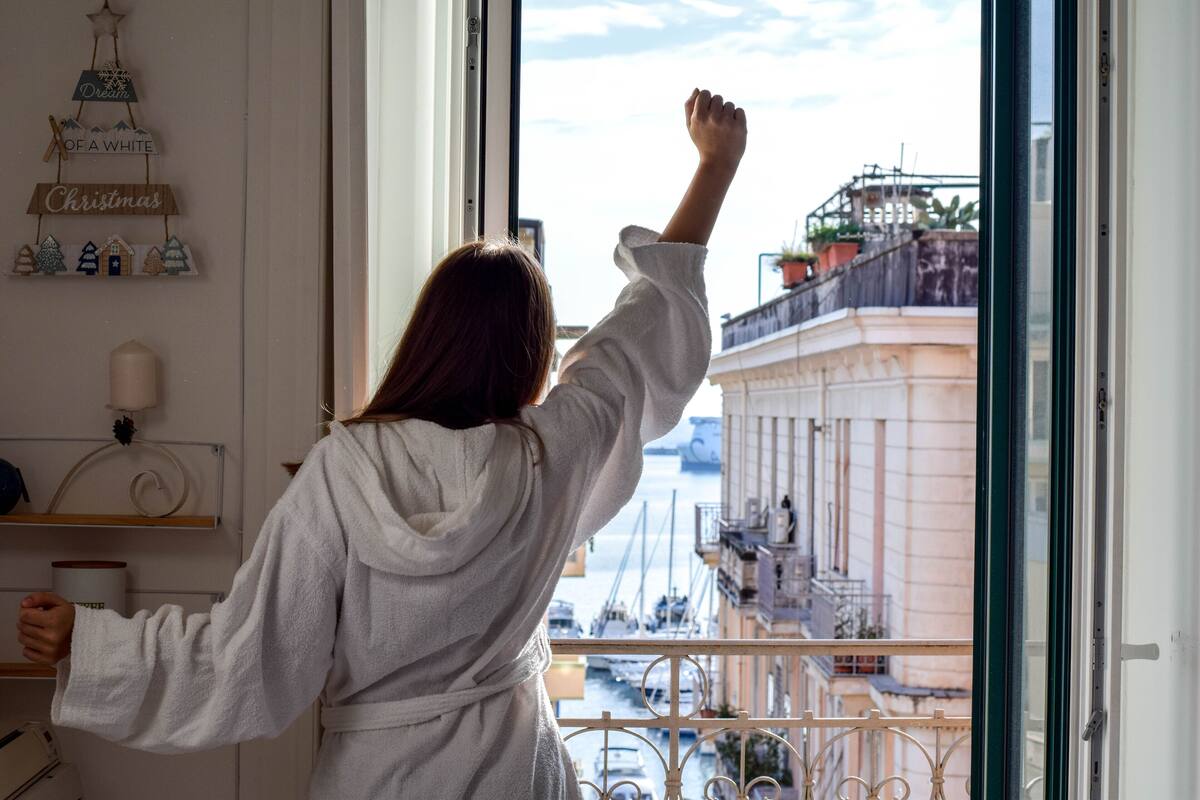 A person in a white robe standing by a window with a view of a harbor and buildings.