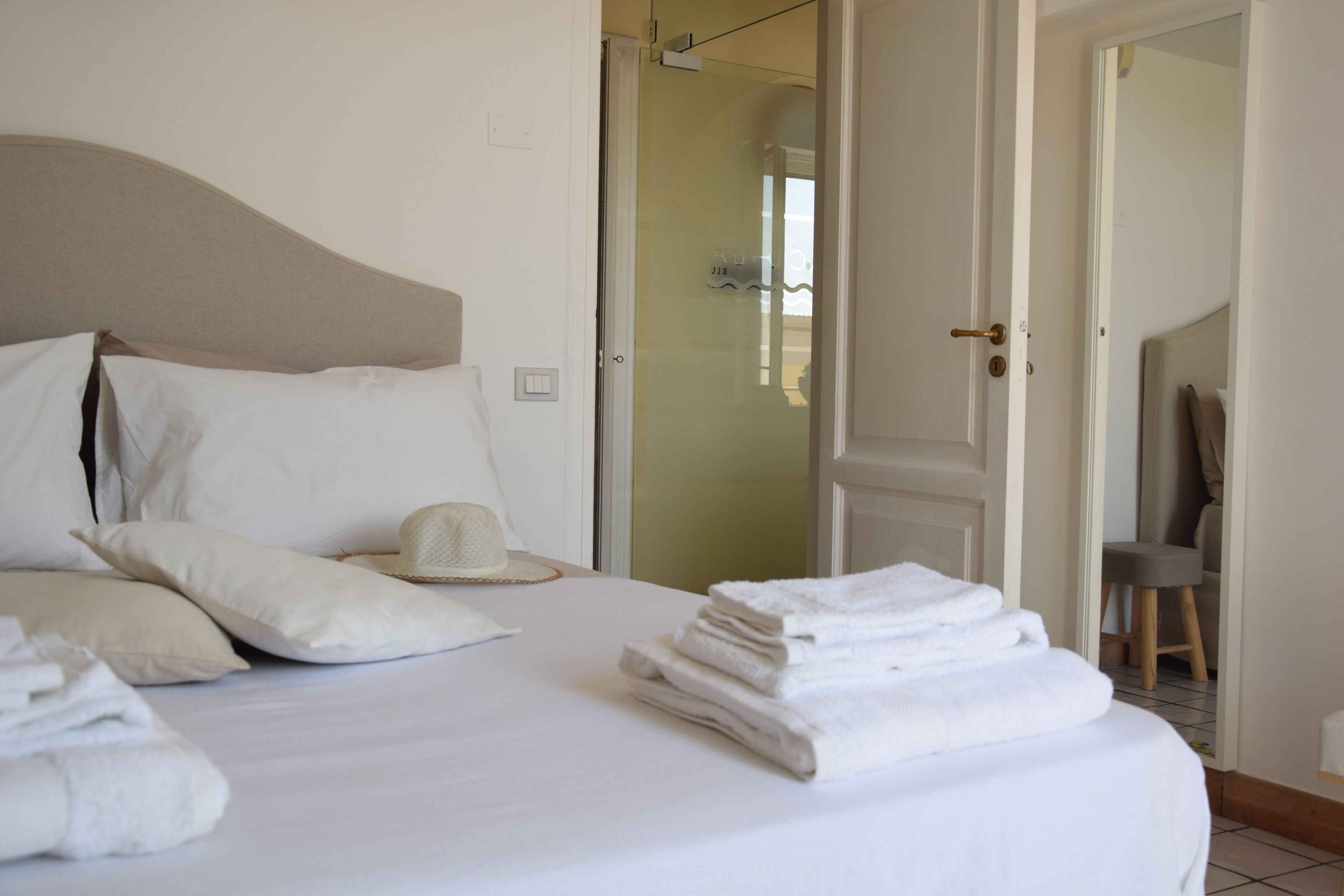 A neatly made bed with pillows and a folded towel, a hat on the bed, and a door leading to another room.