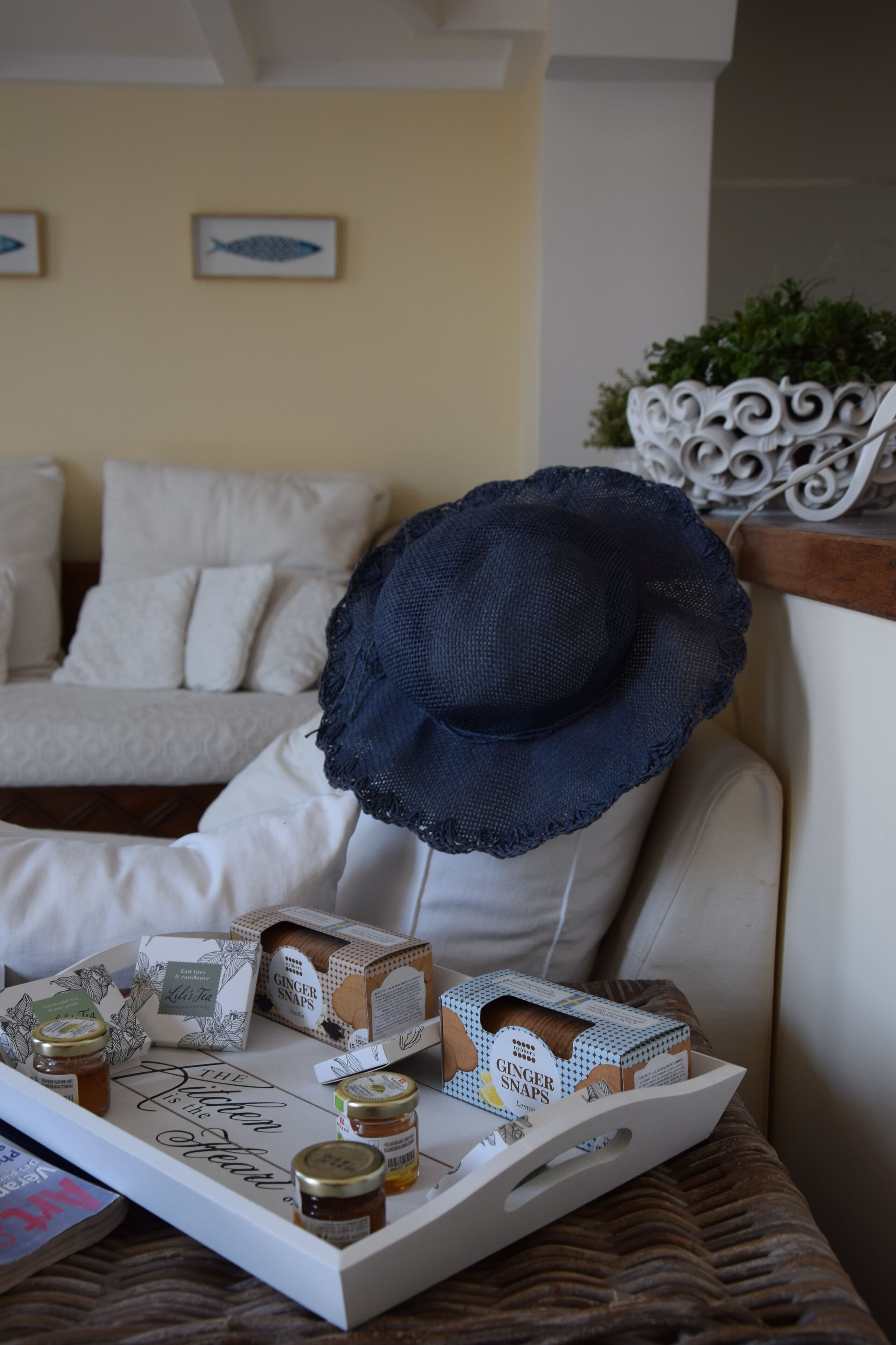 A tray with ginger snaps, tea bags, and jars on a table in a room with a white sofa and decorative fish wall art.