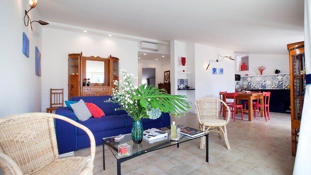 Una sala de estar con un sofá azul, sillas de mimbre, una mesa de centro de vidrio y una zona de comedor con sillas rojas y una mesa de madera.