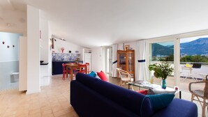 A modern living room with a dark blue sofa, a dining area with red chairs, and a view of mountains through large windows. - Casa Iommart with Terrace (Sant'Agnello)