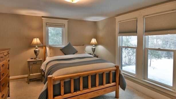 A bedroom with a wooden bed, two bedside tables with lamps, a dresser, and large windows with blinds. - Chequamegon Haus - Comfortable and Charming walk to downtown Cable, WI (Cable)