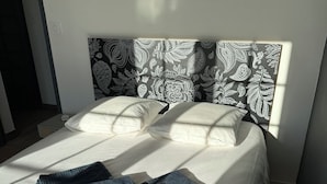 A neatly made bed with white linens and a patterned headboard, a folded pair of dark blue pants on the bed, and a small bedside table with a lamp.