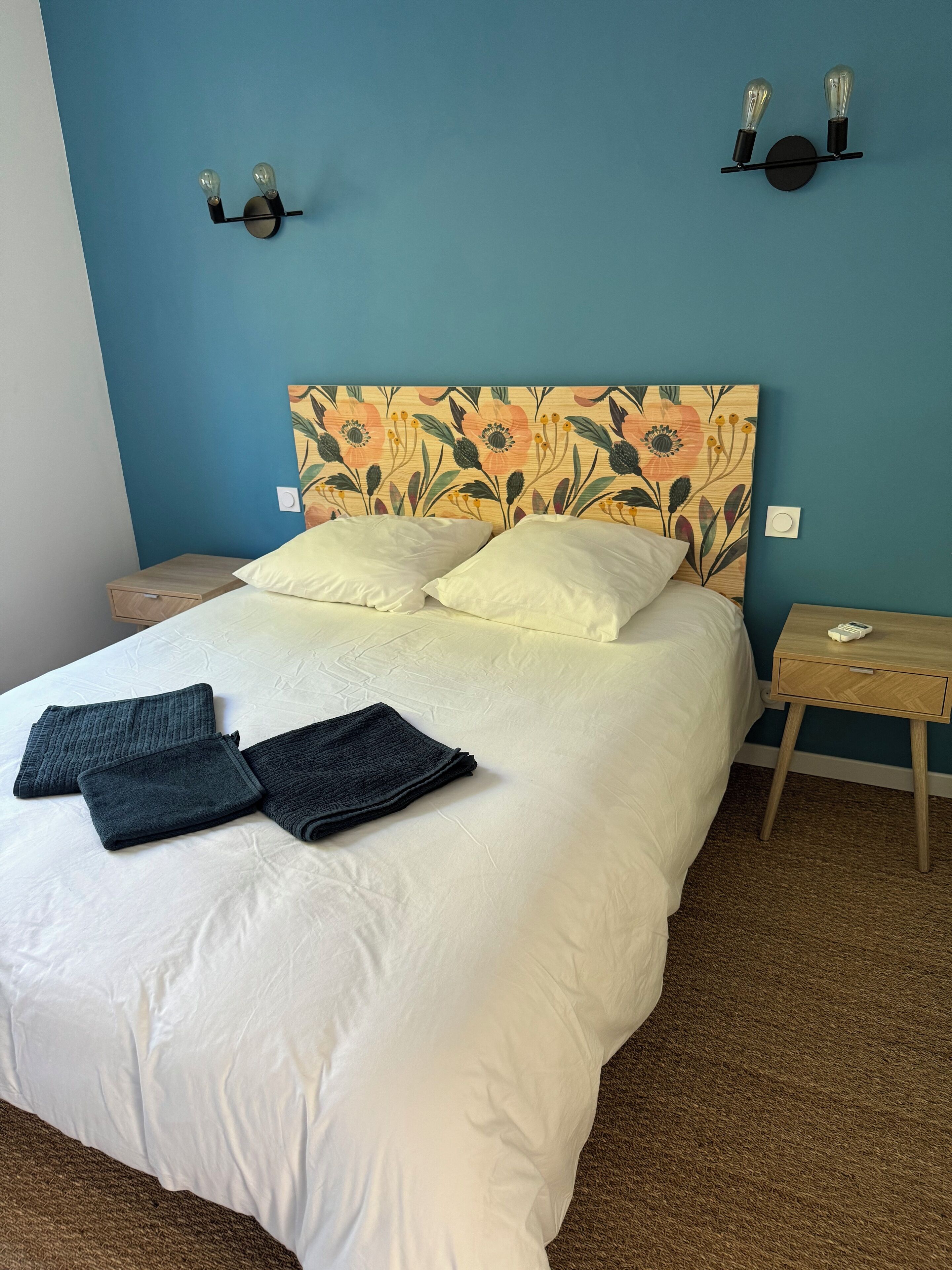 A neatly made bed with pillows and folded blankets, a wooden nightstand with a phone, and a wall-mounted light fixture.