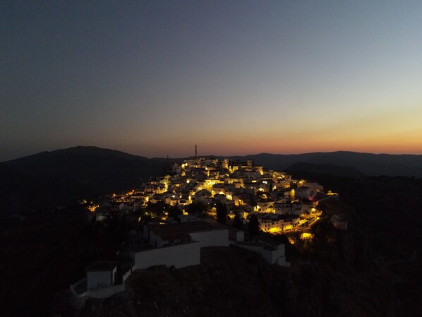 Un pueblo al anochecer con edificios iluminados, rodeado de colinas.