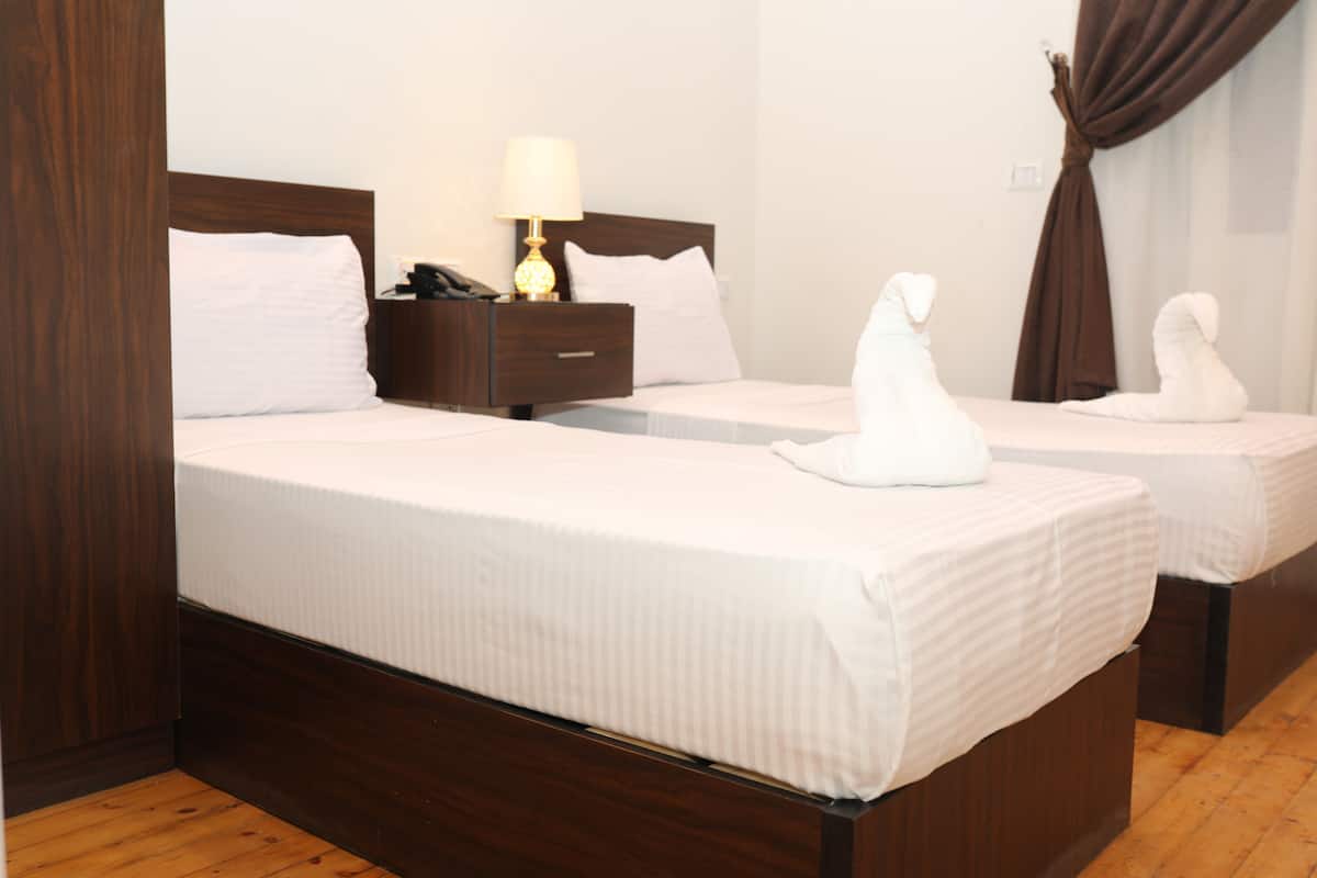 A hotel room with two beds, wooden headboards, white bedding, a nightstand with a lamp, and a curtain.