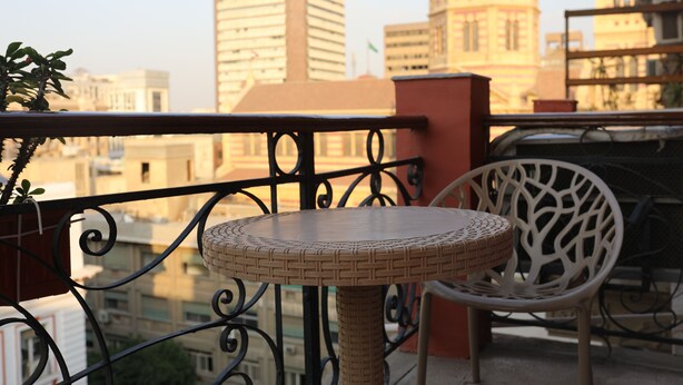 A round wicker table with a metal railing in the foreground, a white chair, and a view of a cityscape with buildings in the background. - New Midtown Hotel (Cairo)