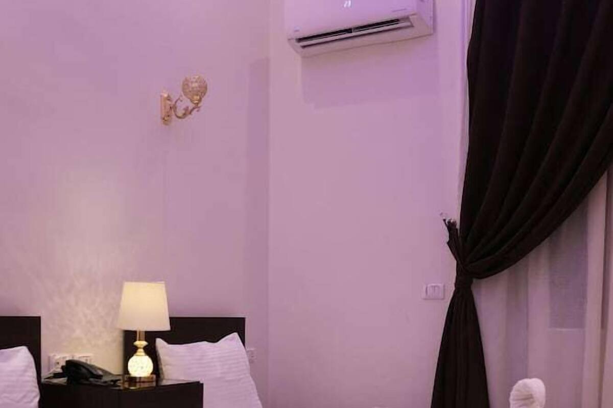 A hotel room with two single beds, a bedside table with a lamp, an air conditioning unit, and a dark curtain.
