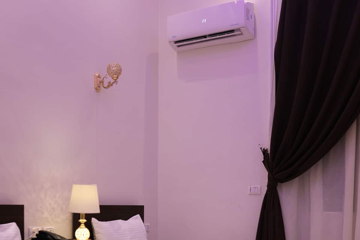 A hotel room with two single beds, a bedside table with a lamp, an air conditioning unit, and a curtain.