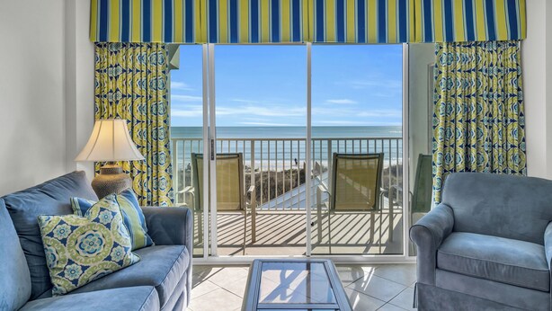 A room with a sofa, armchair, and a table, a balcony with a view of the beach, and patterned curtains. - Oceanfront Condo in Cherry Grove. Enjoy fun in the sun! Reduced Rates! (North Myrtle Beach)