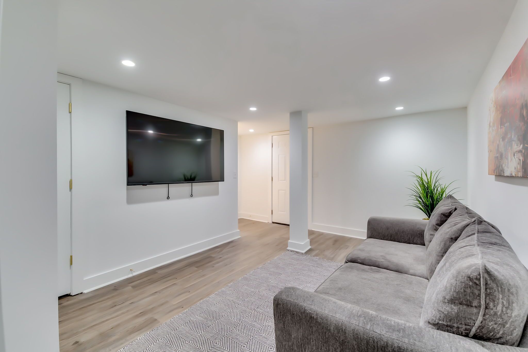 A modern living room with a grey sofa, a patterned rug, a flat-screen TV mounted on the wall, and a plant.