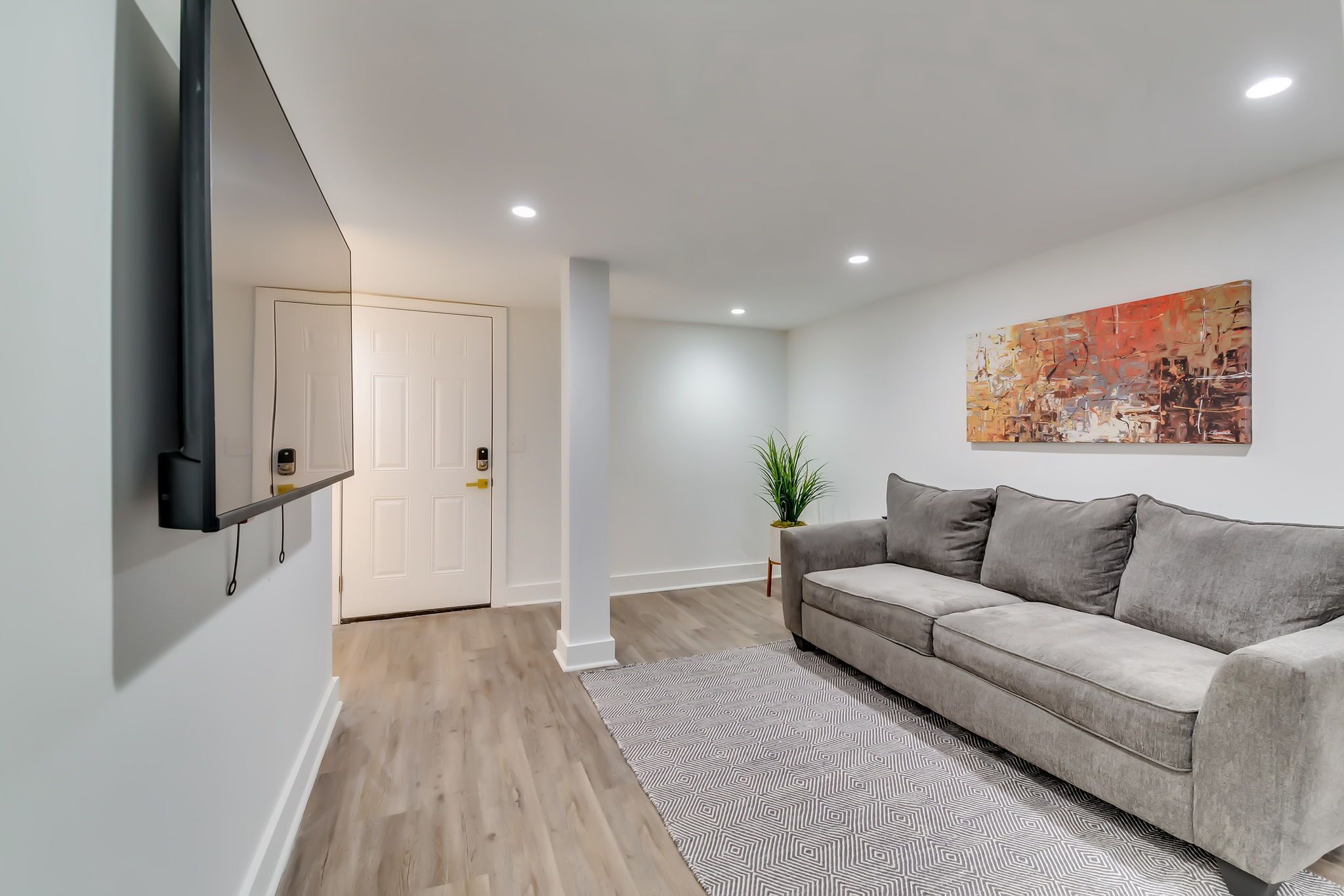 A modern living room with a grey sofa, a patterned rug, a wall-mounted TV, and a door leading to another room.