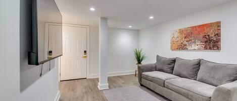A modern living room with a grey sofa, a patterned rug, a wall-mounted TV, and a door leading to another room.