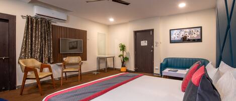 A hotel room with a bed, a desk, a chair, a sofa, a ceiling fan, an air conditioner, and a potted plant.