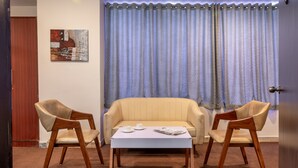 Une pièce avec un canapé beige, deux chaises en bois, une petite table blanche sur laquelle se trouvent des tasses et un magazine, et un tableau accroché au mur.