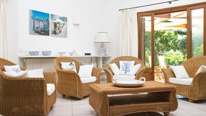 A room with wicker furniture, a white tiled floor, a table with a tray, a lamp, and two paintings on the wall.