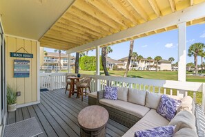 A covered outdoor area with a sofa, chairs, and a table. There are signs on the wall, including one that says 'BEACH HOUSE' and another with advice from the ocean.