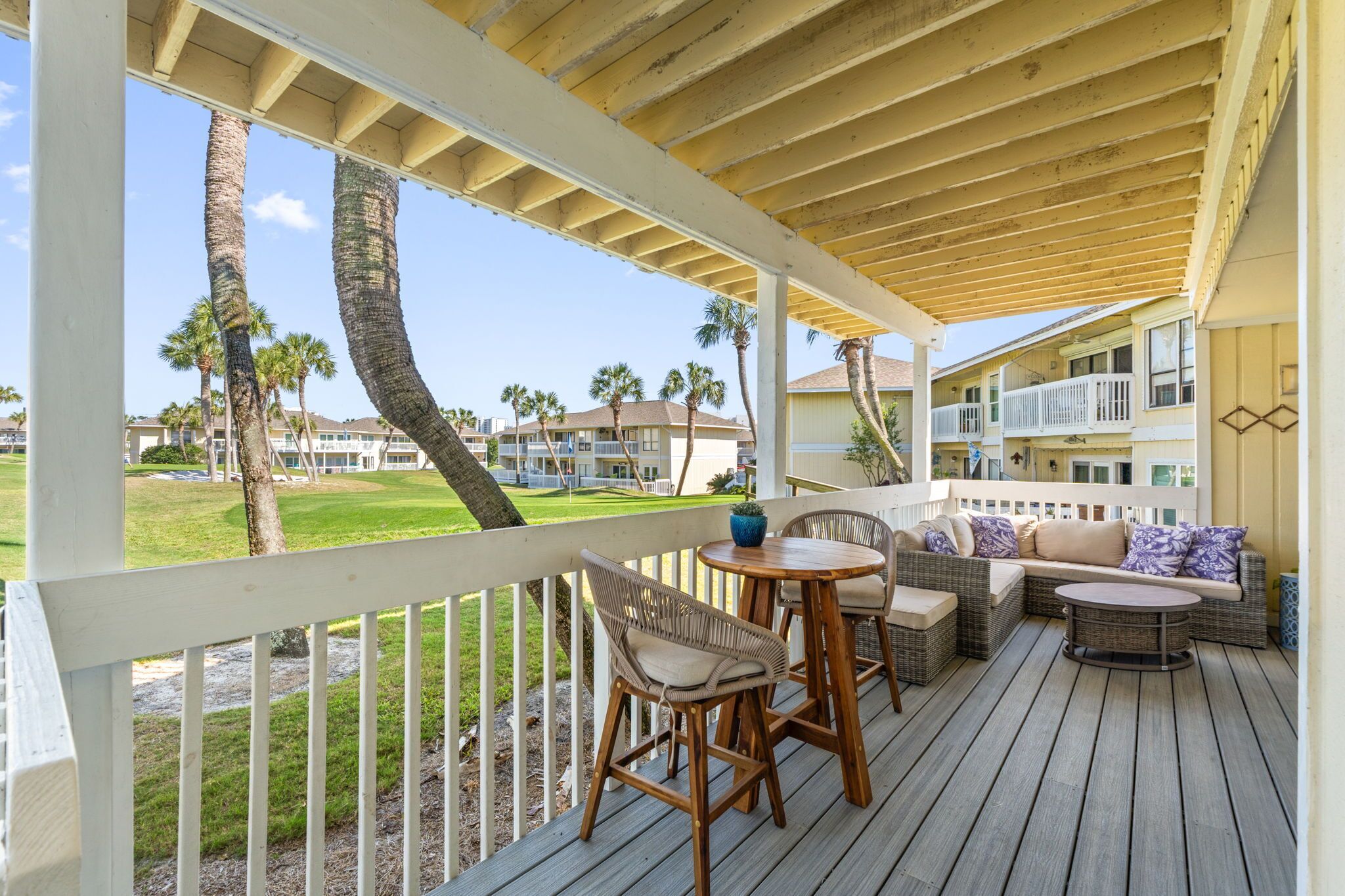 Un espacio exterior cubierto con una terraza de madera, muebles de mimbre y vistas a un campo de golf y edificios.