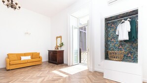 A room with a herringbone floor, a mustard-colored sofa, a wooden cabinet, and a wardrobe with a tropical wallpaper. - Lux Aurea (Meta)