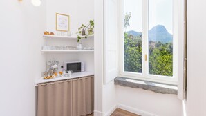 A small kitchen with white cabinets, a microwave, and a window with a view of greenery and a mountain. - Lux Aurea (Meta)