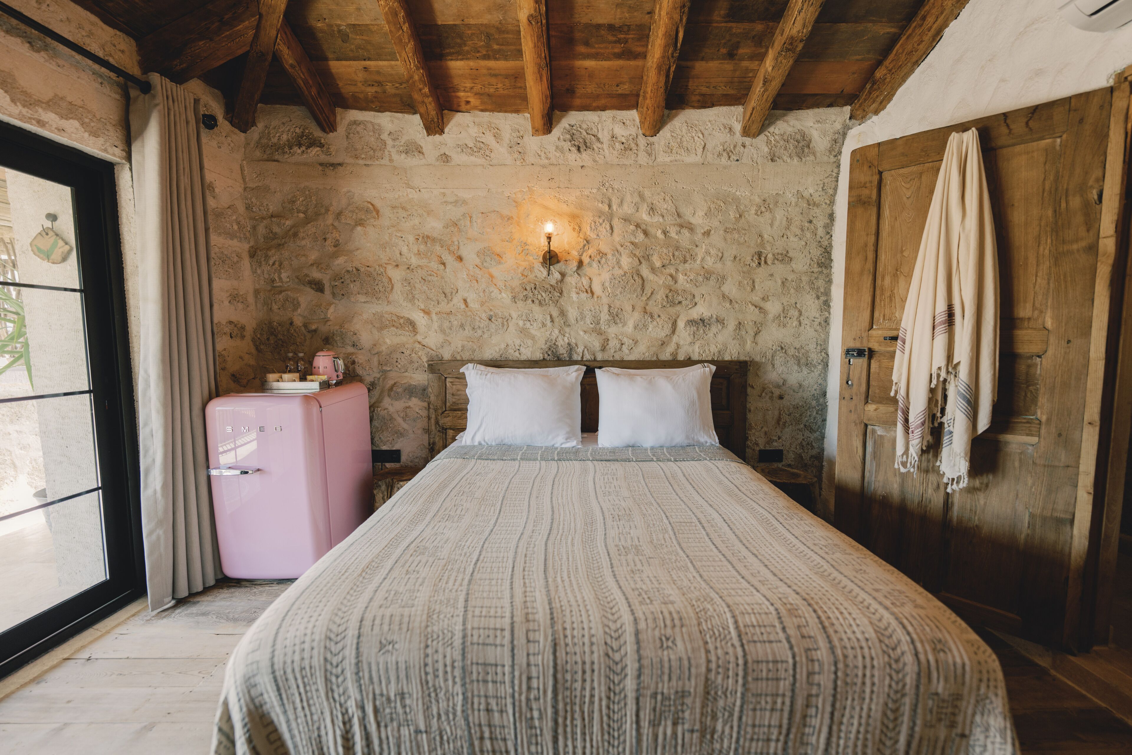 A rustic bedroom with a stone wall, wooden ceiling beams, a bed with a patterned bedspread, a pink vintage refrigerator, and a wooden door with a towel hanging on it.