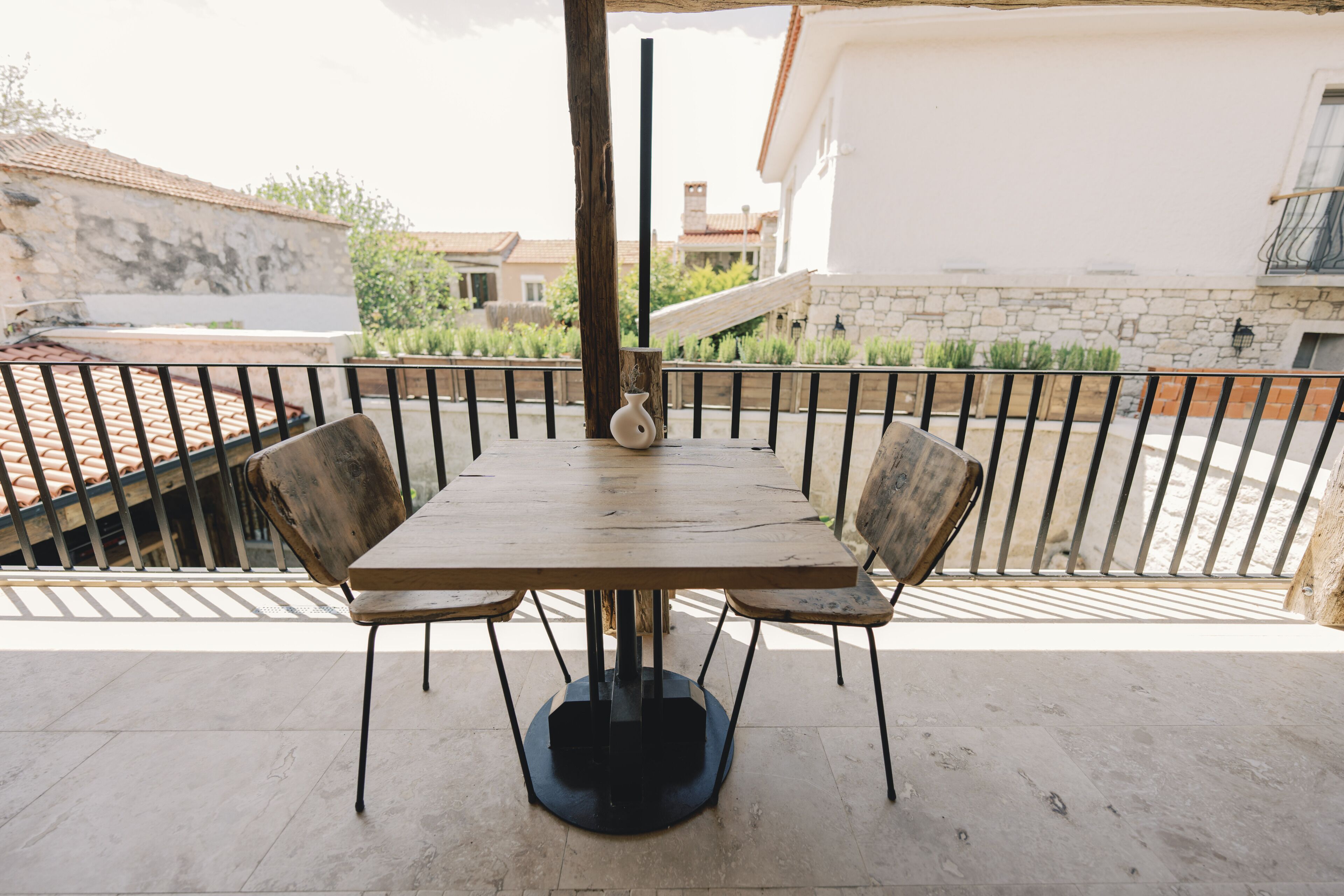 Une table en bois, entourée de quatre chaises, sur un balcon. En arrière-plan, on aperçoit un mur en pierre et une balustrade.