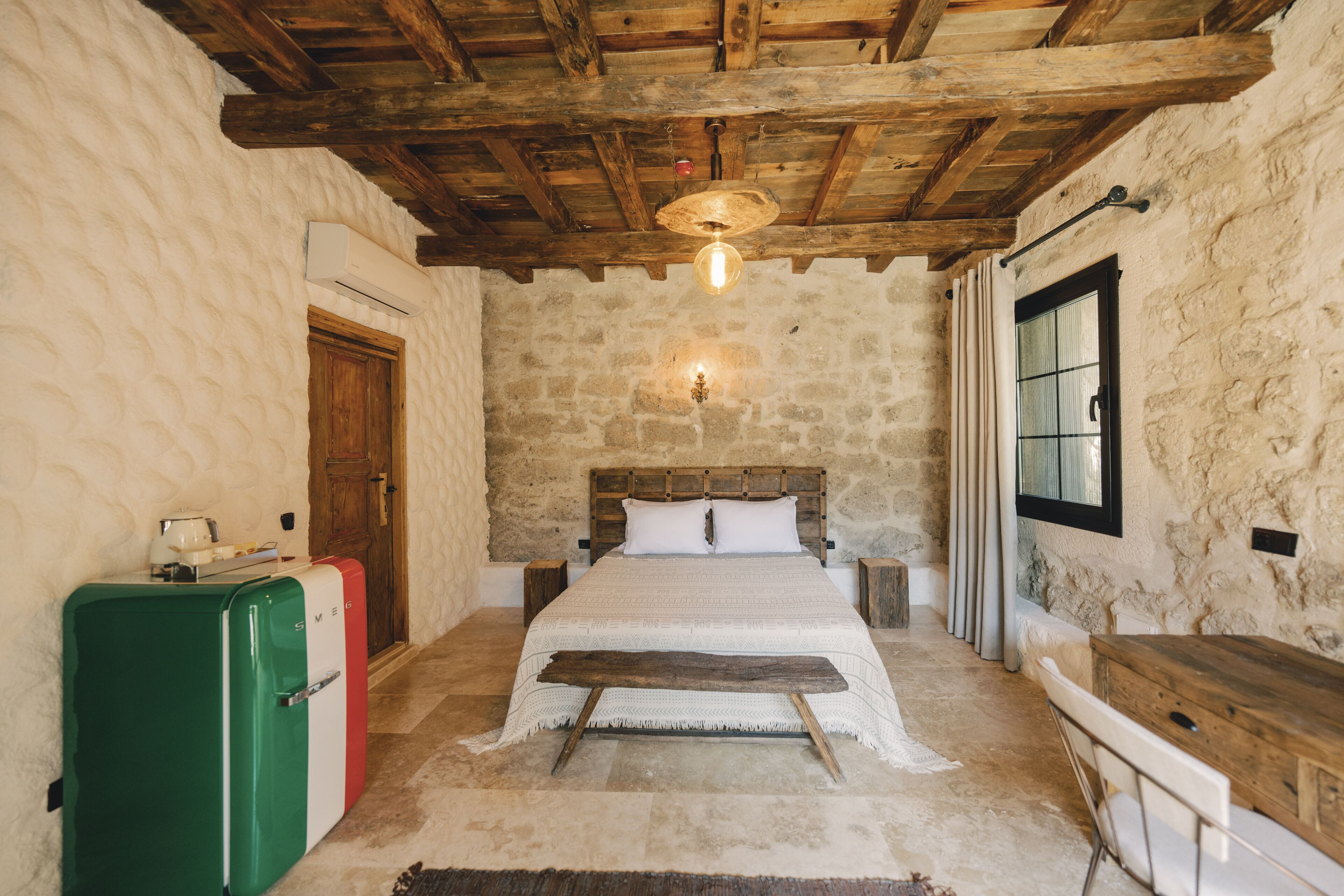 A rustic bedroom with a stone wall, wooden ceiling beams, a bed with a wooden headboard, a small table, and a vintage-style refrigerator.