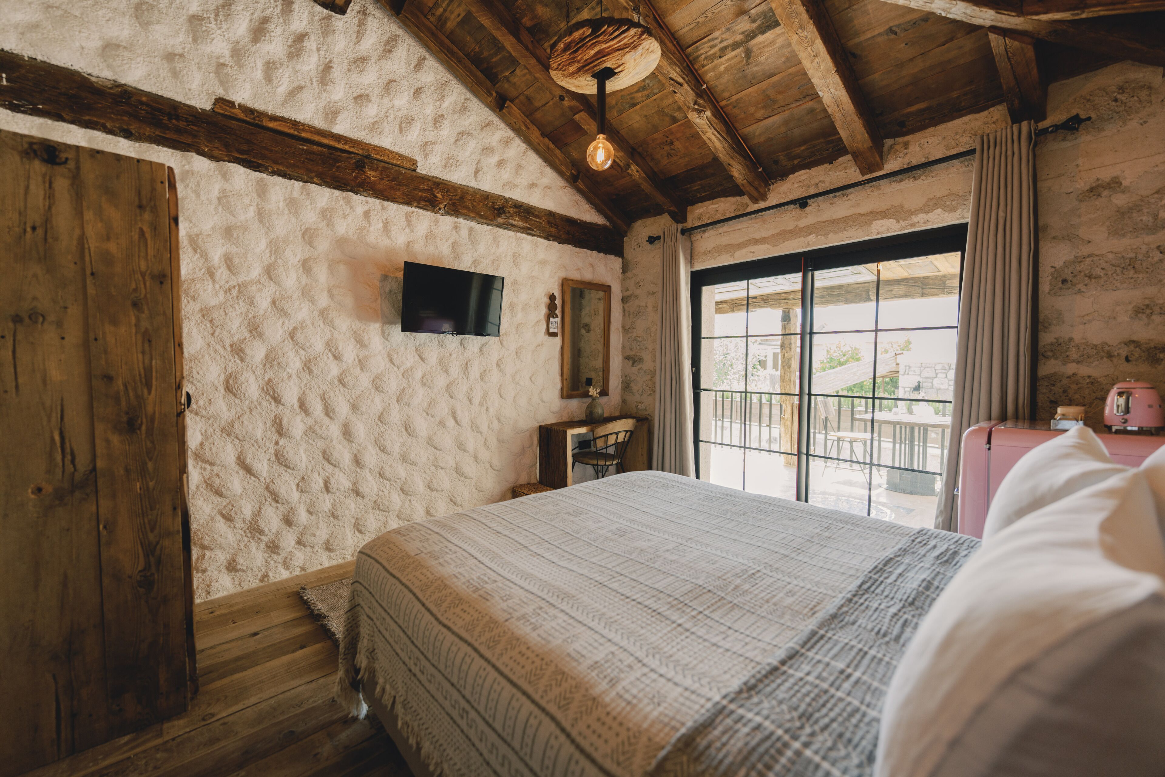 A rustic room with a bed, wooden beams, a television, and a window with curtains.