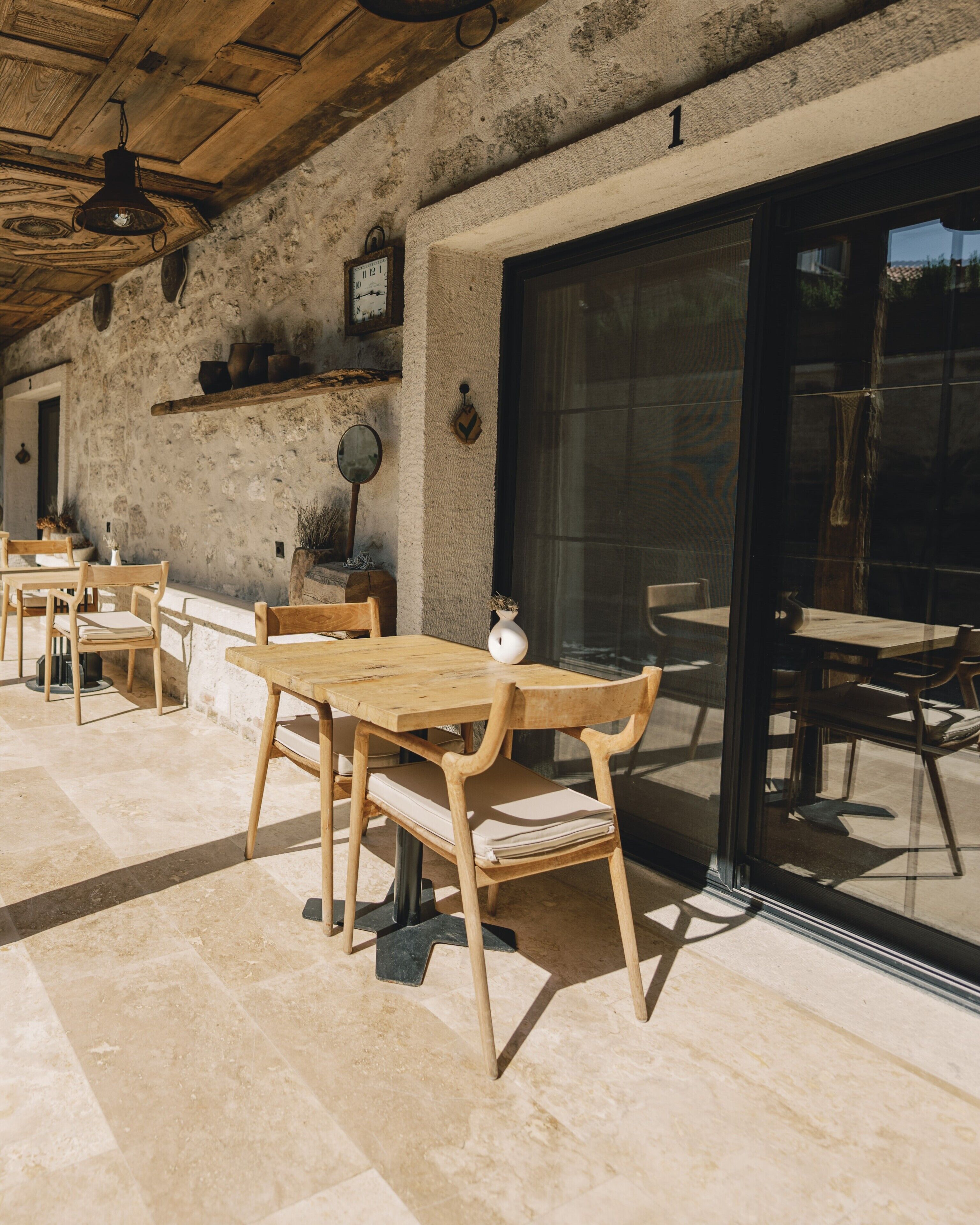 A rustic outdoor dining area with wooden tables and chairs, stone walls, and a large sliding glass door.
