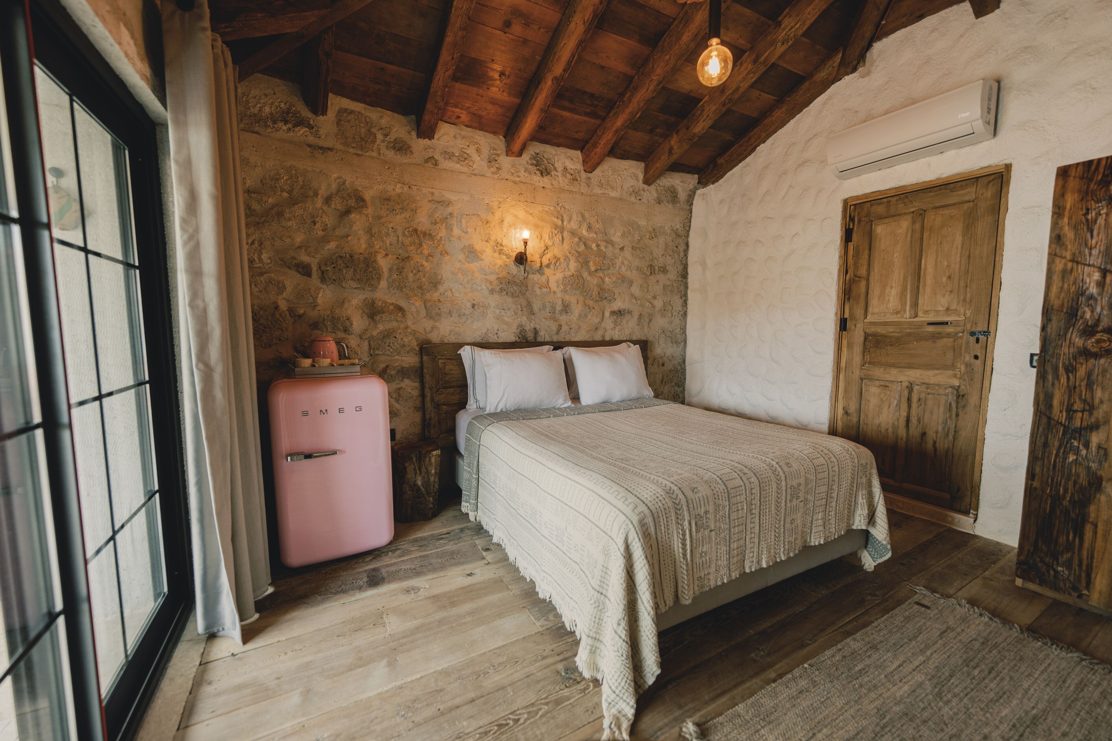 A rustic bedroom with a stone wall, wooden ceiling beams, a bed with a white bedspread, a pink suitcase, and a wooden door.