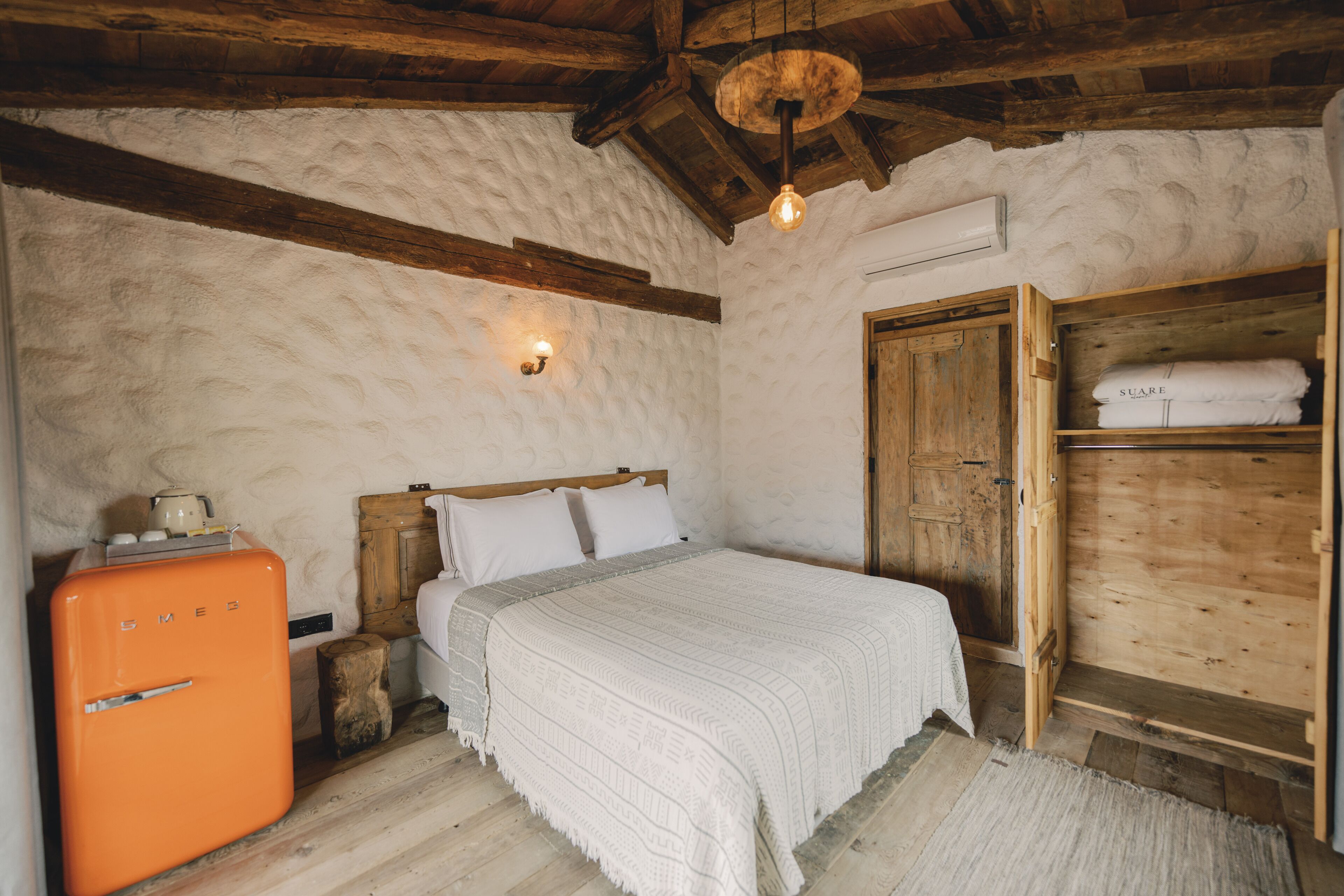A rustic bedroom with a wooden bed, a vintage orange refrigerator, and a wooden wardrobe.